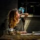 Woman with long wavy hair sitting at a rustic table, gently touching her neck as if experiencing tension or pain, with a teapot and cup beside her — symbolizing the role of mindful nutrition and anti-inflammatory foods in chronic pain relief, FL licensed dietitian nutritionist